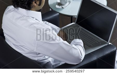 Mideastern Man Using Laptop At Office, With A Cup Of Cofee And Documents On The Side Table