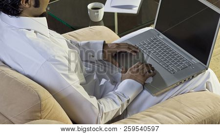 Mideastern Man Using Laptop At Office, With A Cup Of Cofee And Documents On The Side Table