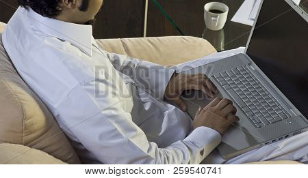 Mideastern Man Using Laptop At Office, With A Cup Of Cofee And Documents On The Side Table
