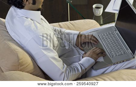 Mideastern Man Using Laptop At Office, With A Cup Of Cofee And Documents On The Side Table