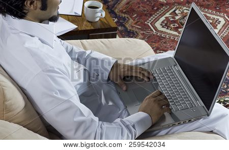 Mideastern Man Using Laptop At Office, With A Cup Of Cofee And Documents On The Side Table