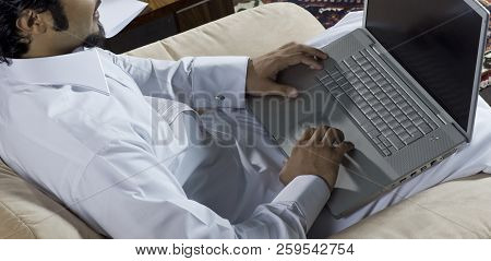 Mideastern Man Using Laptop At Office, With A Cup Of Cofee And Documents On The Side Table