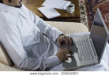 Mideastern Man Using Laptop At Office, With A Cup Of Cofee And Documents On The Side Table