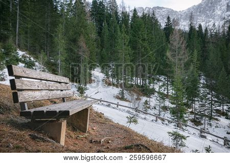 Early Winter Landscape With A Wooden Bench On The Side Of An Alpine Road, Snowy Fir Forest And Mount