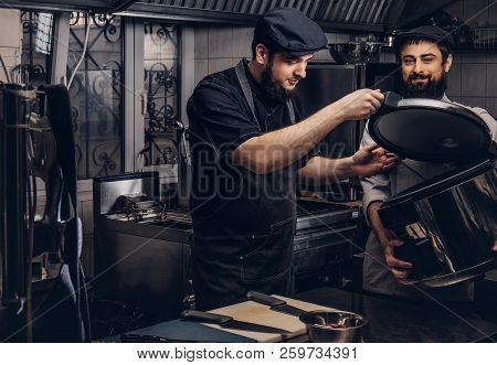 Two Brutal Cooks Dressed In Uniforms And Hats Standing With Big Multicooker In The Kitchen.