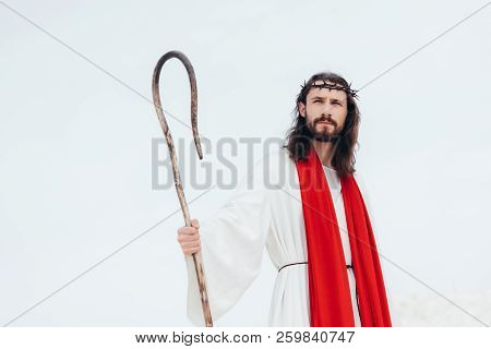 Low Angle View Of Jesus In Robe, Red Sash And Crown Of Thorns Standing With Wooden Staff In Desert