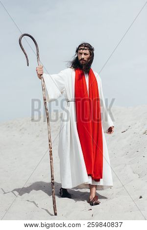 Jesus In Robe, Red Sash And Crown Of Thorns Standing With Wooden Staff In Desert And Looking Away