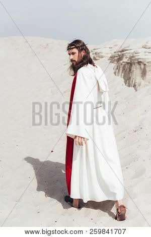 Back View Of Jesus In Robe, Red Sash And Crown Of Thorns Walking In Desert