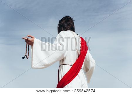 Back View Of Jesus In Robe, Red Sash And Crown Of Thorns Holding Rosary Against Blue Sky