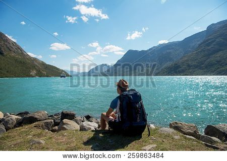 Hiker With Backpack Sitting And Looking At Gjende Lake In Jotunheimen National Park, Norway