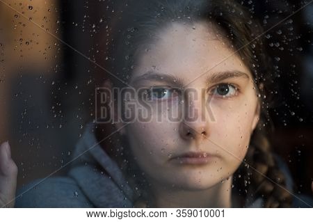 Young Girl Is Looking With Hope The Window On Which Raindrops, Is Staying At Home Isolation For Stav
