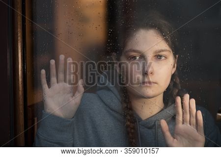 Young Girl Is Looking With Hope The Window On Which Raindrops, Is Staying At Home Isolation For Stav
