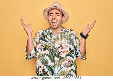 Young handsome man with blue eyes on vacation wearing summer florar shirt and hat celebrating mad and crazy for success with arms raised and closed eyes screaming excited. Winner concept
