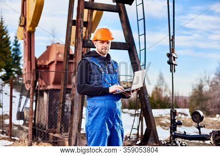 Portrait Of Petrolium Engineer, Wearing Blue Overalls And Orange Helmet, With A Laptop, Standing Wit