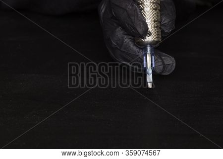 Part Of A Rotary Tattoo Machine With A Needle In Hand In A Black Glove On A Black Background. Side V