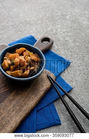 Bowl With Deicious Kimchi On Wooden Cutting Board And Blue Cloth Near Chopsticks On Concrete Surface