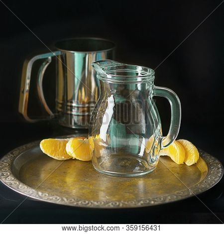 Glass Decaner And Metal Mug And Several Bright Segments Of Orange On Vintage Tray On Dark Background