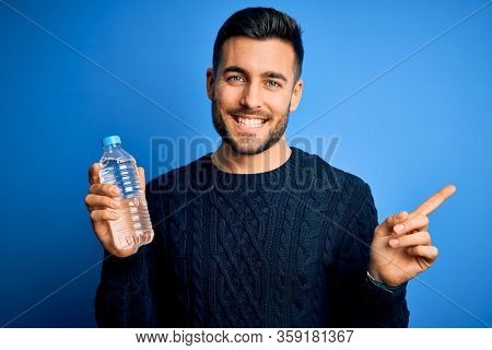Young handsome man drinking bottle of water to refeshment over blue background very happy pointing with hand and finger to the side