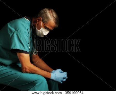 Exhausted Middle Aged doctor in scrubs, gloves and mask sitting with head down amid the Covid-19 crisis, profile portrait against black.