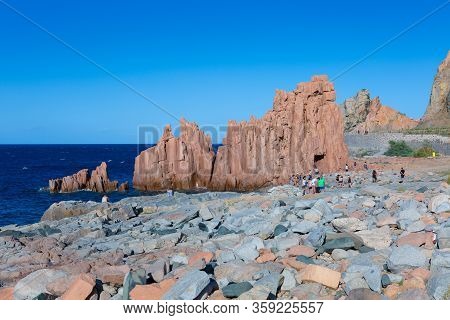 Sardegna, Italy - July 2019: Sardinia Coastline: Typical Red Rocks And Cliffs And Tourists Near Sea 