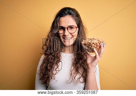 Beautiful woman with curly hair holding bowl with healthy peanuts over yellow background with a happy face standing and smiling with a confident smile showing teeth
