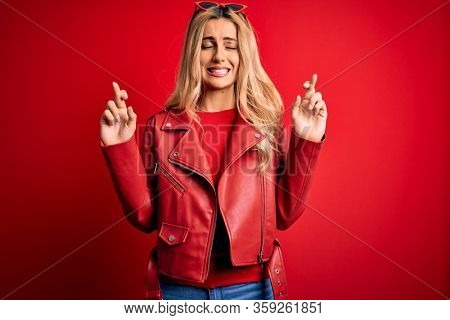 Young beautiful blonde woman wearing casual jacket standing over isolated red background gesturing finger crossed smiling with hope and eyes closed. Luck and superstitious concept.