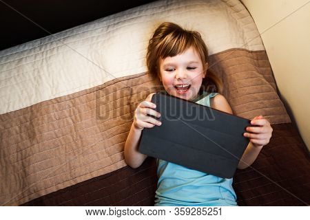 Todder Girl Laying On The Bed And Holding Tablet, Looking At It And Smiling