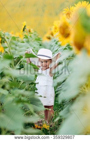 A Cute Little Smiling Girl In The Field Of Sunflowers. Child In Momy Hat. Childhood Concept. Sunny S