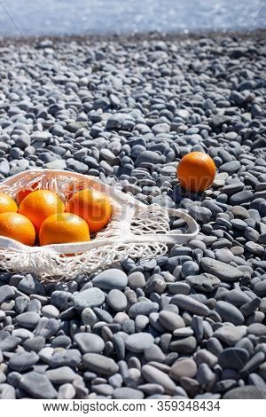 White Cotton Eco String Bag With Oranges On A Rocky Beach. Sea View With A Blue Sky. Zero And Eco Wa