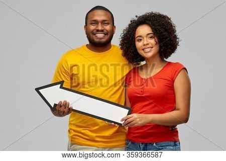 direction and people concept - happy smiling african american couple holding big leftwards thick arrow over grey background