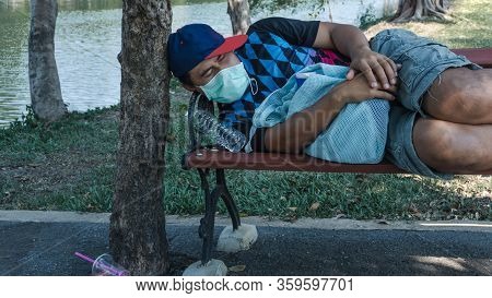 Asian Homeless Man Wearing Protective Mask Sleeping On A Bench. Unemployed Due To The Impact Of The 