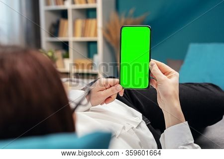 Back View Of Person Holding Vertical Cellphone With Green Screen Lying On Bed Or Sofa In Apartment