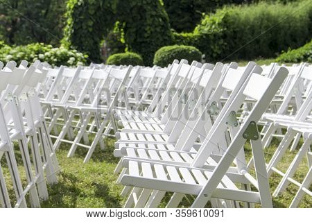 View Of Empty Wooden Chairs In White, Arranged In Rows Against A Background Of Park Plants, Close-up
