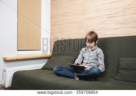 Lonely boy sittng on sofa with tablet computer stuck at home during isolation. Quarantine and lockdown are protective measures against spreading of coronavirus pandemic disease