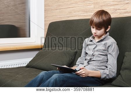 Lonely boy sittng on sofa with tablet computer stuck at home during isolation. Quarantine and lockdown are protective measures against spreading of coronavirus pandemic disease