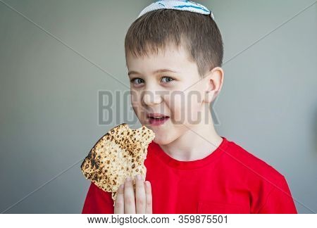 Cute Caucasian Child In A White Kippah Cap Eating Shmura Matzo, A Piece Of Traditional Jewish Unleav