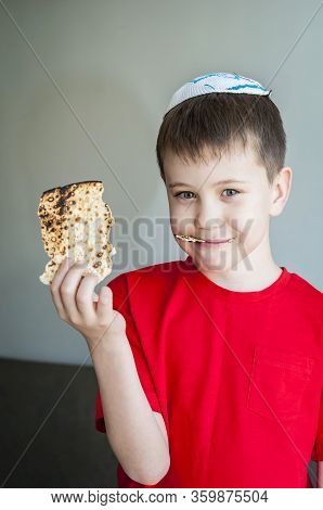 Cute Caucasian Child In A White Kippah Cap Eating Shmura Matzo, A Piece Of Traditional Jewish Unleav