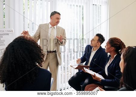 Serious Mature Entrerpreneur Answering Questions Of Young Colleagues At Meeting