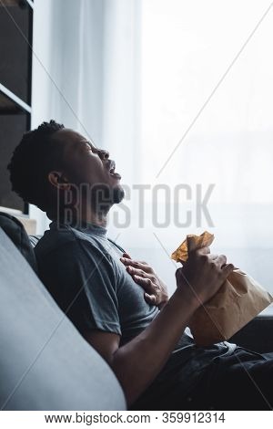 Stressed African American Man Holding Paper Bag While Having Panic Attack At Home