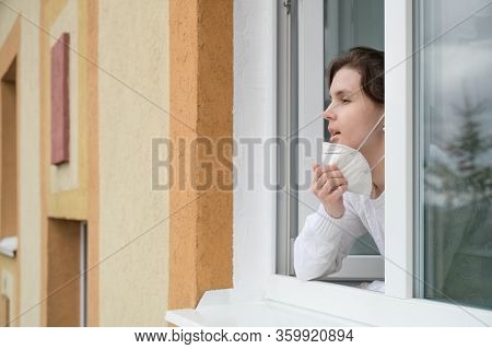 Caucasian Young Woman Smiles, Looks Out Of The Apartment Window And Takes Off Her Respirator. End Of