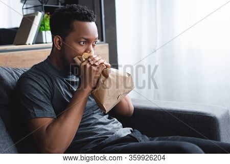 Stressed African American Man Breathing With Paper Bag While Having Panic Attack At Home