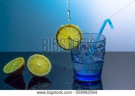 Splashes Of Water Fall Into A Glass With Lemon And Ice.a Mug Of Ice And Water Sits On A Mirrored Tab