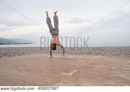 Shirtless Caucasian Man Doing Backflip On Pebble Beach.