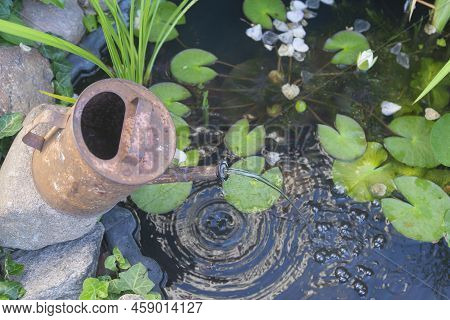 An Old, Rusty Watering Can In The Form Of A Fountain On An Artificial Pond.
