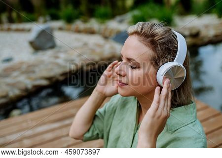 Rlaxed Woman Wearing Headphones Listening To Music Sitting On A Pier By Natureal Lake In Summer