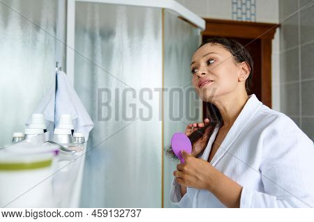 Charming Woman Looking At Her Reflection In The Bathroom Mirror And Brushing Hair With Comb, Caring 