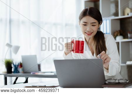 An Asian Female Employee Sits With A Red Coffee Mug In The Startup's Marketing Department Office, Sh