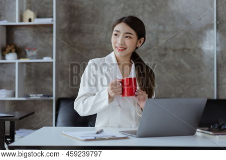 An Asian Female Employee Sits With A Red Coffee Mug In The Startup's Marketing Department Office, Sh