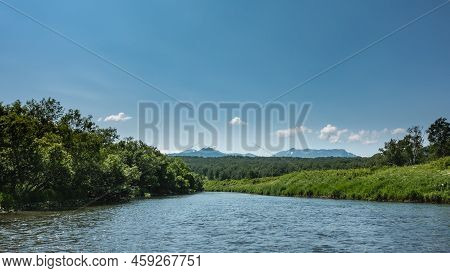 The Bed Of The Blue River Bends Between Banks Overgrown With Lush Green Vegetation. A Mountain Range