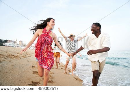 Happy Group Of Young People Having Fun At The Beach. Cheerful Friends Run Holding Hands Enjoying The